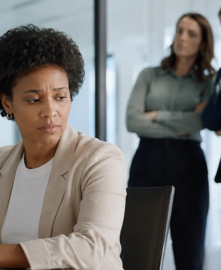 Employee sits alone in a modern office while two colleagues watch from the background, suggesting workplace exclusion and discrimination.