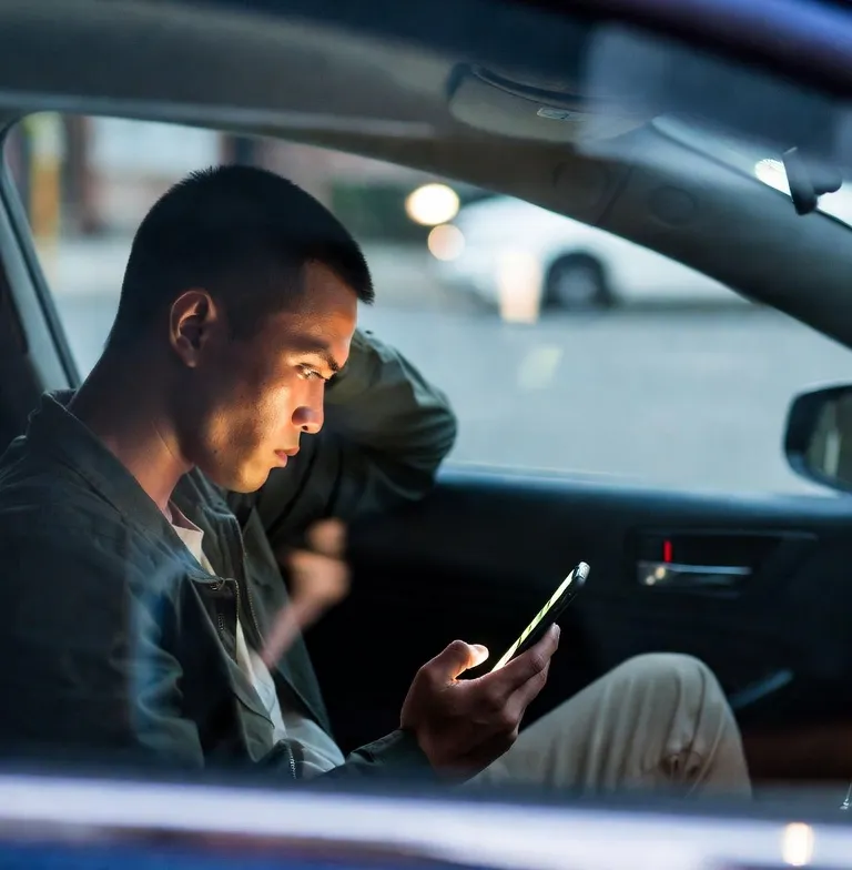 A concerned man sitting inside a parked car at dusk, intently checking his glowing smartphone, with soft evening street lights visible through the windows.