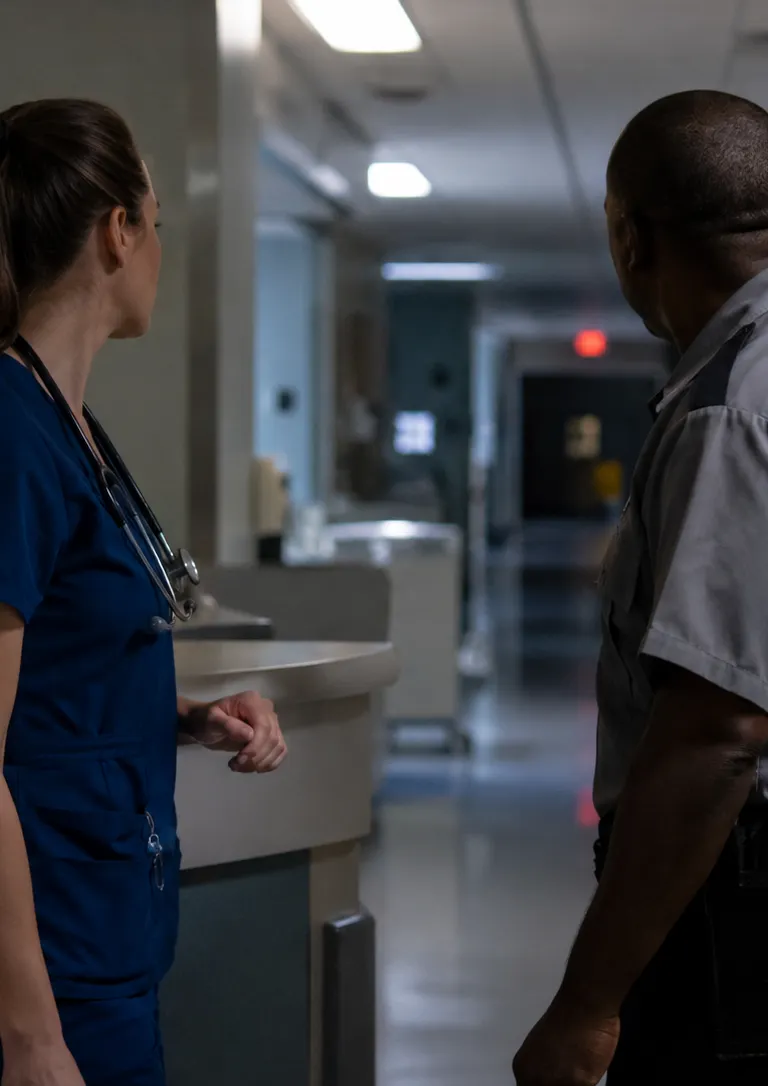 Nurse in scrubs standing near a nurses' station at night speaking with a uniformed security guard, both looking toward an unseen disturbance down a hospital corridor.