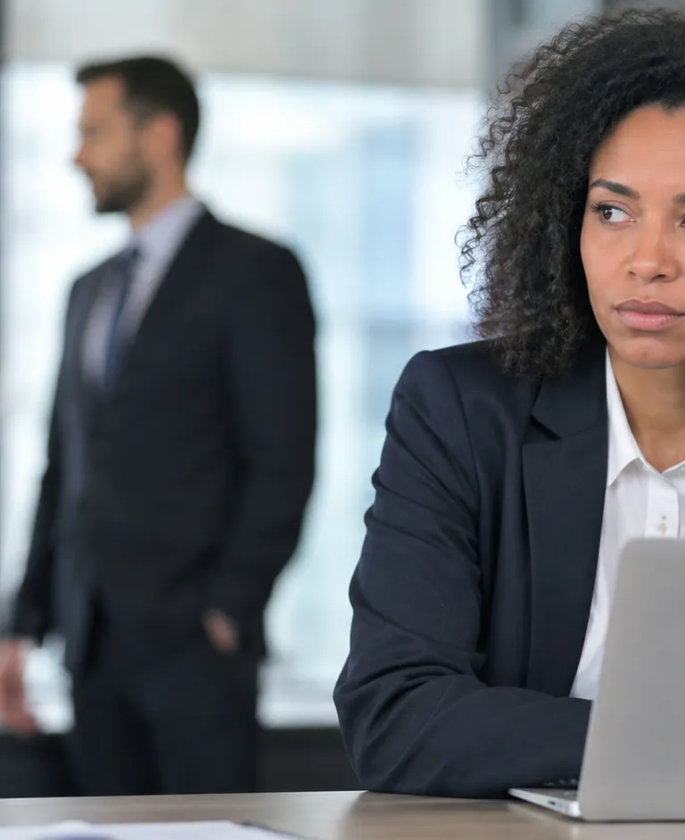 Pensive office worker sitting at a laptop while two colleagues talk in the background, illustrating workplace pay disparity concerns.